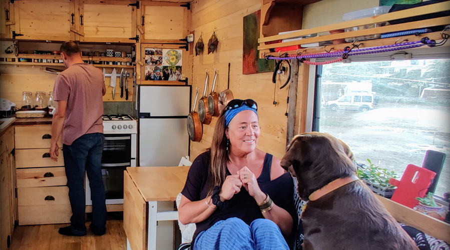 a man cooking inside his mobile home, a woman sitting on a chair with their dog