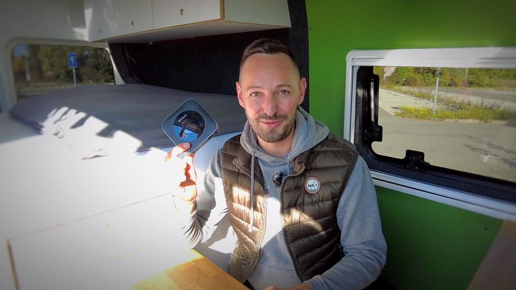 Ein Mann sitzt am Tisch in einem Campervan und hält einen kleinen Wlan-Router in die Höhe.