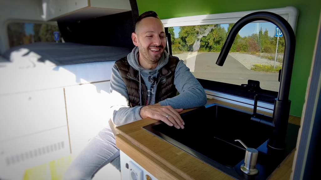 Ein Mann sitzt in seinem Campervan und präsentiert sein Spülbecken.